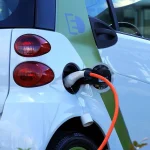 A driver stands between a plug-in hybrid SUV and an EV with a home charger visible in natural light.