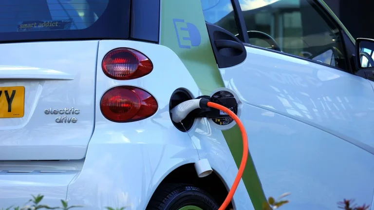 A driver stands between a plug-in hybrid SUV and an EV with a home charger visible in natural light.